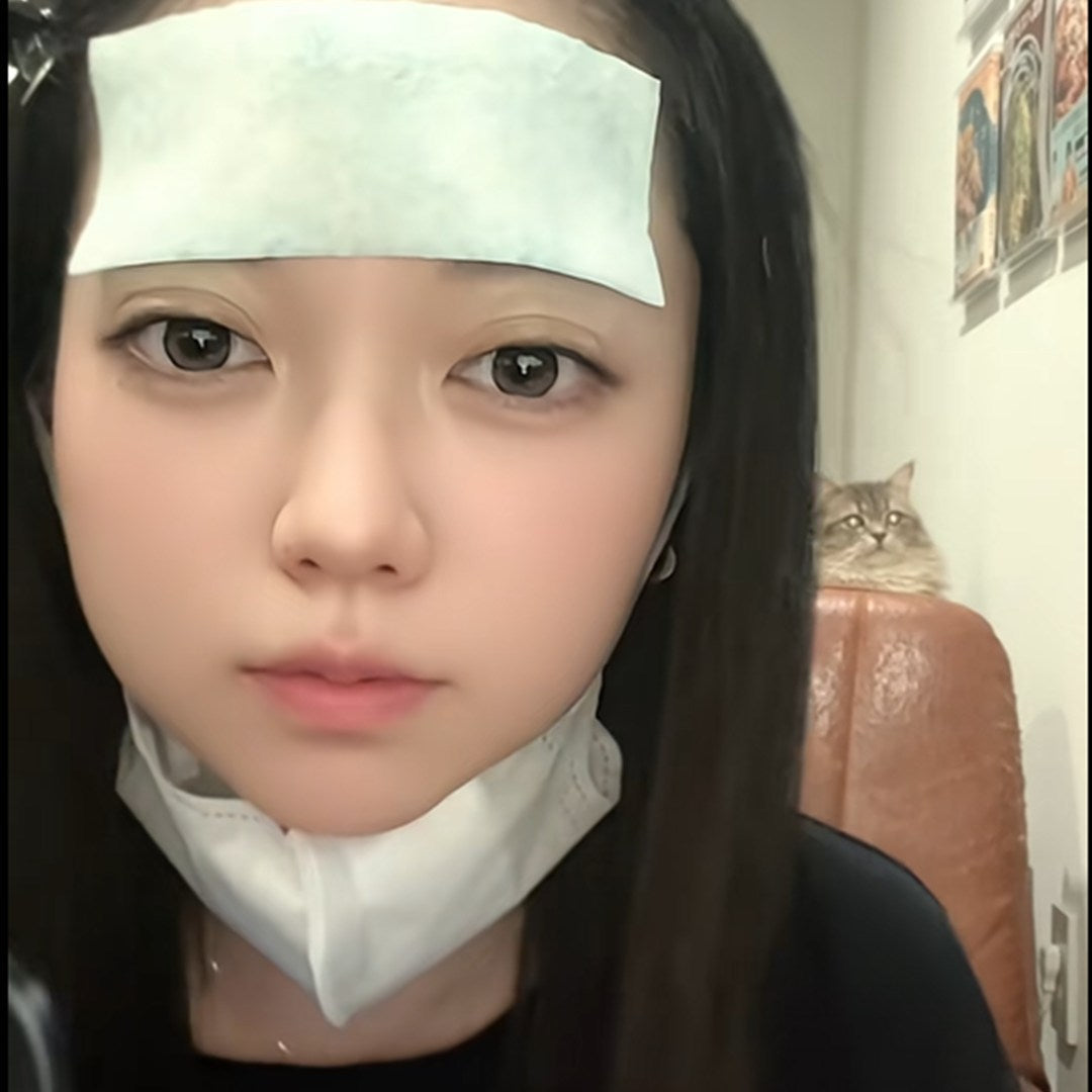おでこに冷却シートを貼り、マスクをあごにかけたマリナ。後ろの椅子から愛猫のぺちが見ている